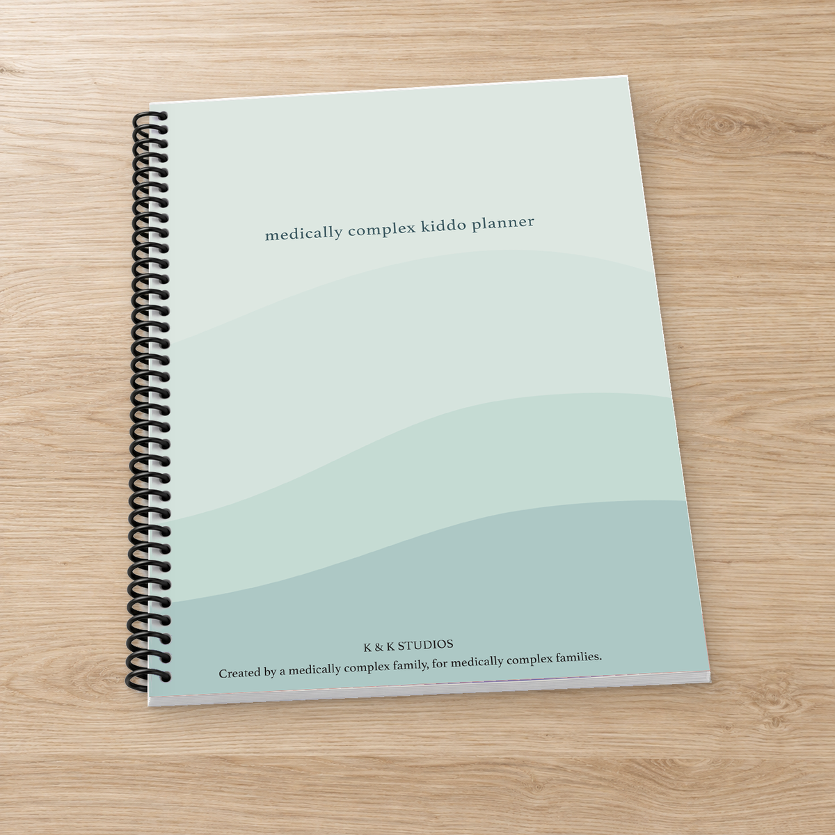A spiral-bound notebook titled ' medically complex kiddo planner' lies on a light wood surface. The cover has a soft mint and pale blue wavy design with black spiral binding and small text at the bottom reading K&K Studios.